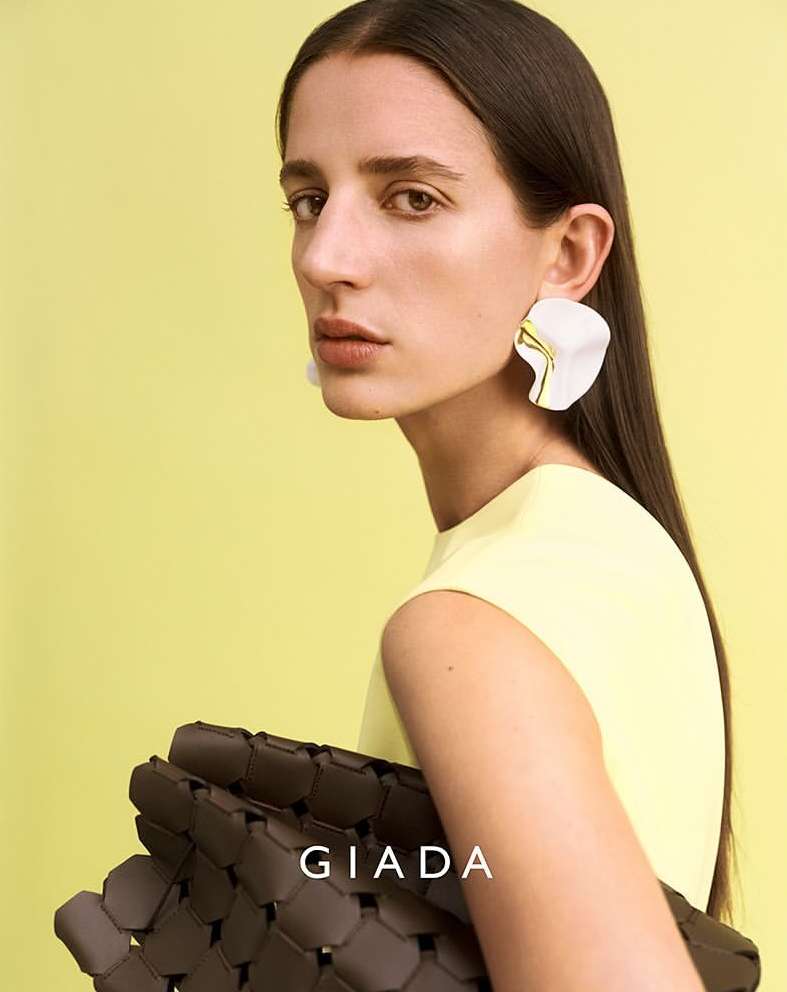 Fashion shot of a woman wearing statement earrings and a yellow top, holding a woven brown purse, against a yellow background.