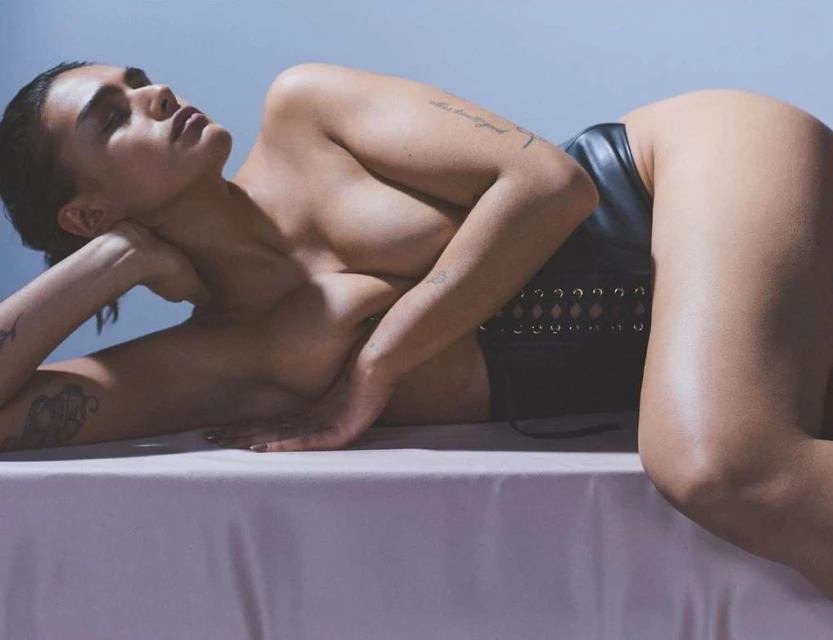Artistic nude portrait of a fair-skinned adult female lying on a light pink surface in a black corset with light blue backdrop.