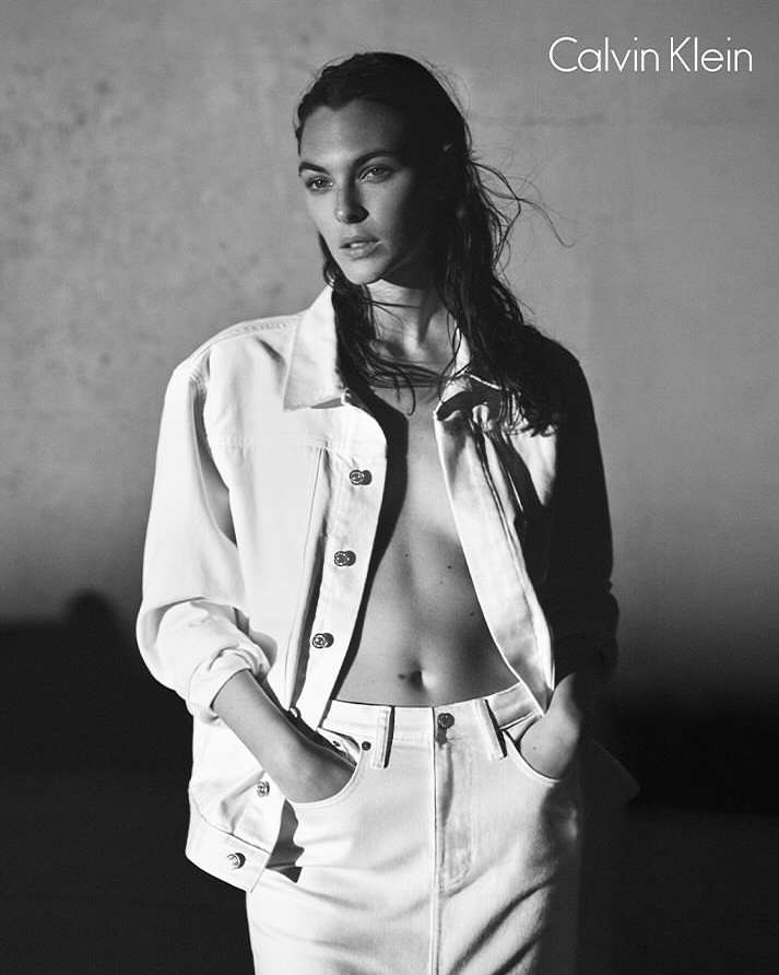 Monochrome fashion shot of a woman in an unbuttoned white denim jacket and matching jeans, standing in front of a dark gray background.