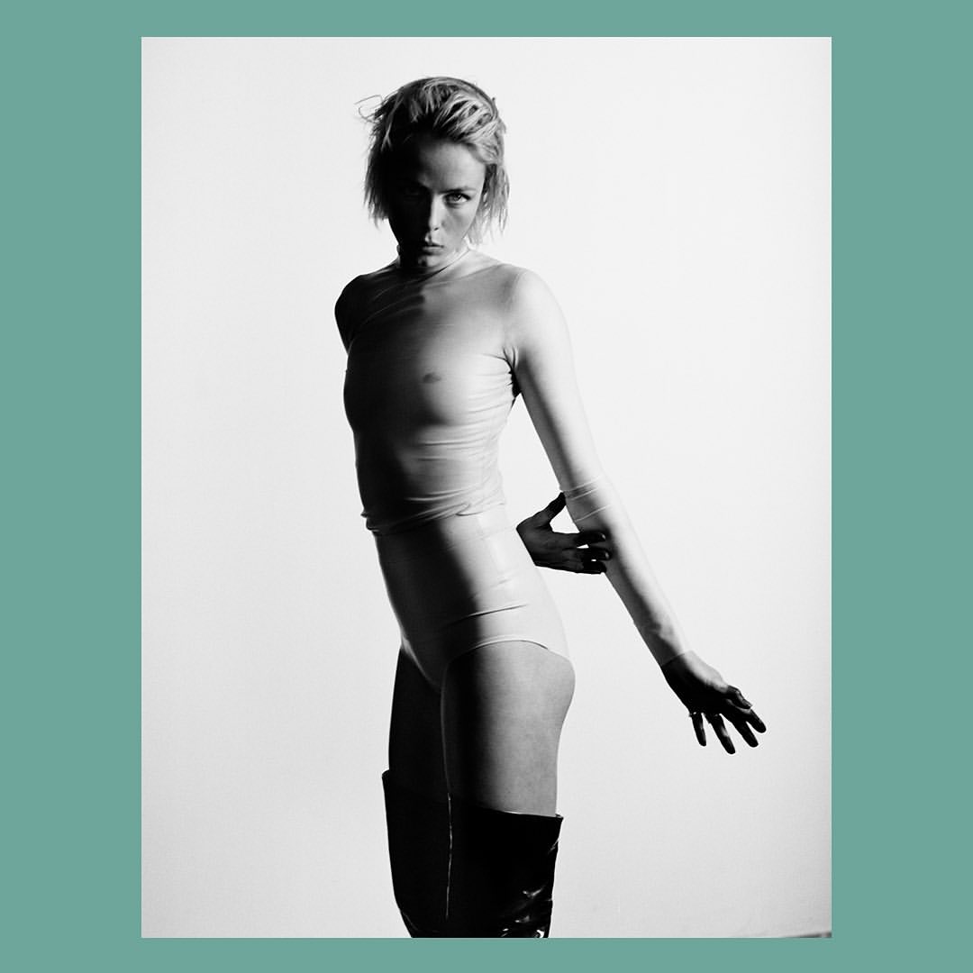 Black and white studio shot of a woman in a sheer top, leotard, and knee-high boots, posing with her arm extended against a white background.