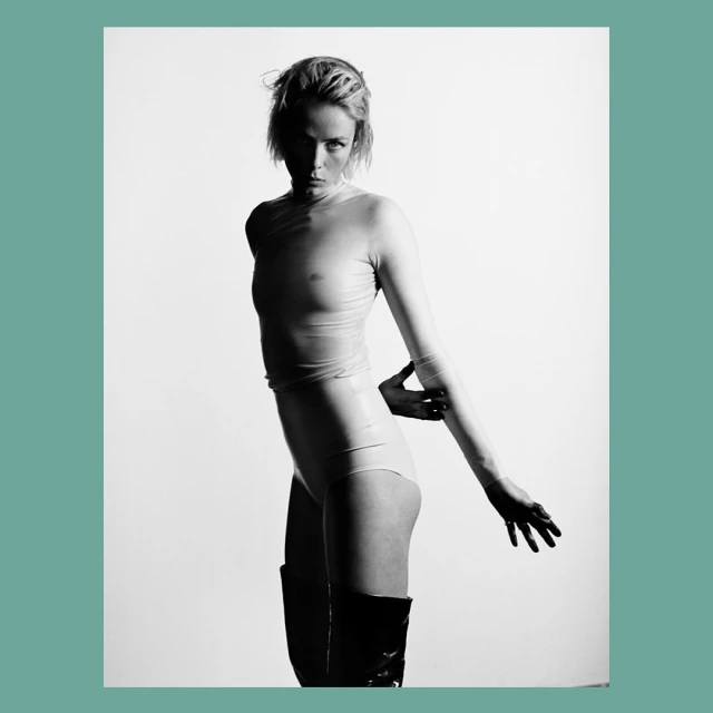Black and white studio shot of a woman in a sheer top, leotard, and knee-high boots, posing with her arm extended against a white background.
