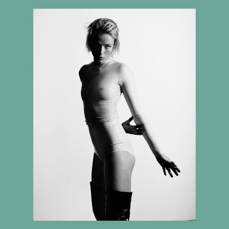 Black and white studio shot of a woman in a sheer top, leotard, and knee-high boots, posing with her arm extended against a white background.