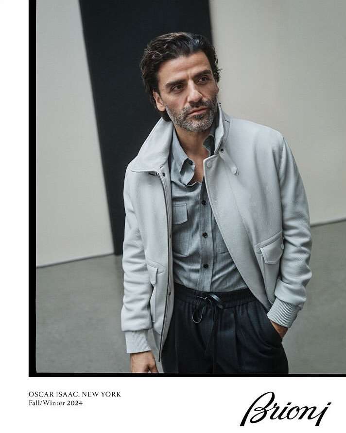 Actor Oscar Isaac poses in a fashion editorial wearing a light gray jacket, grey shirt, and black pants against a neutral background.