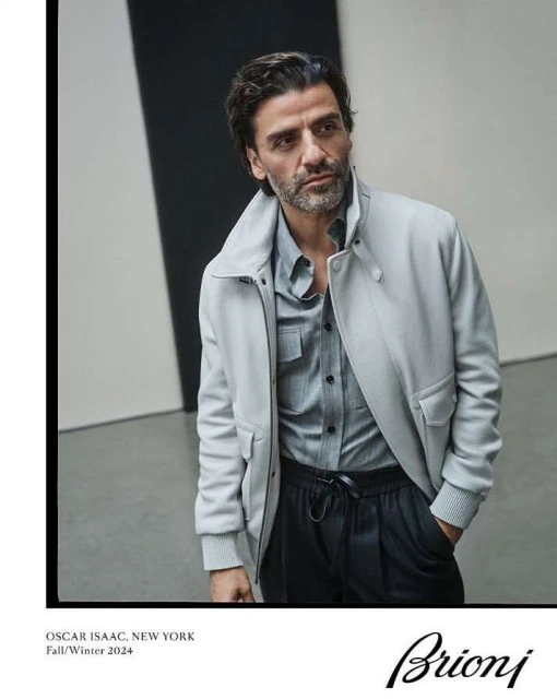 Actor Oscar Isaac poses in a fashion editorial wearing a light gray jacket, grey shirt, and black pants against a neutral background.
