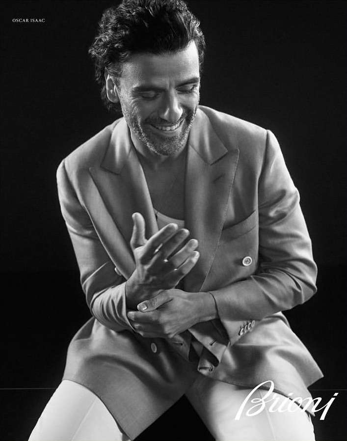 Oscar Isaac sits smiling in a light-colored suit and white pants against a dark background in a portrait-style studio image.