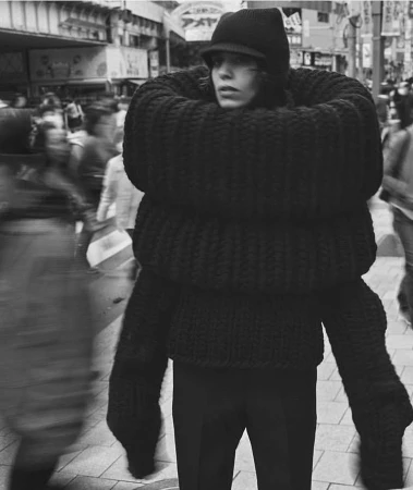 Person in oversized black knitwear stands on city street surrounded by blurred pedestrians, creating a surreal, high-fashion editorial image.