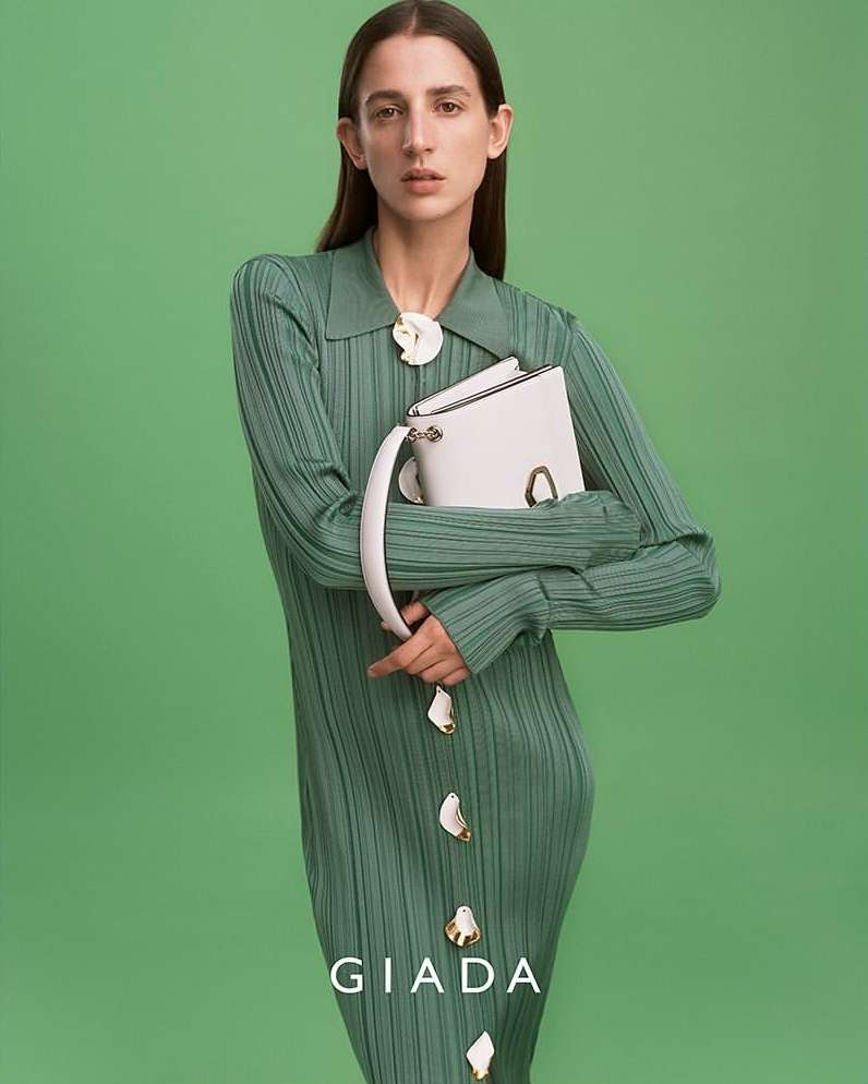 A female model presents a white handbag in a studio setting, wearing a seafoam green dress with matching flower accents and a green backdrop.