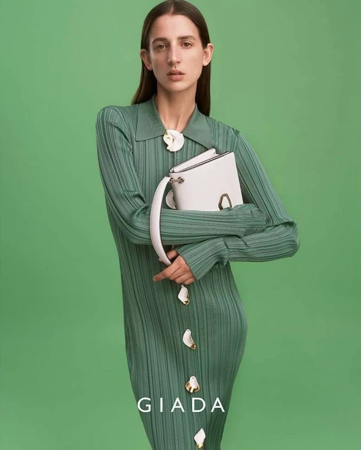 A female model presents a white handbag in a studio setting, wearing a seafoam green dress with matching flower accents and a green backdrop.