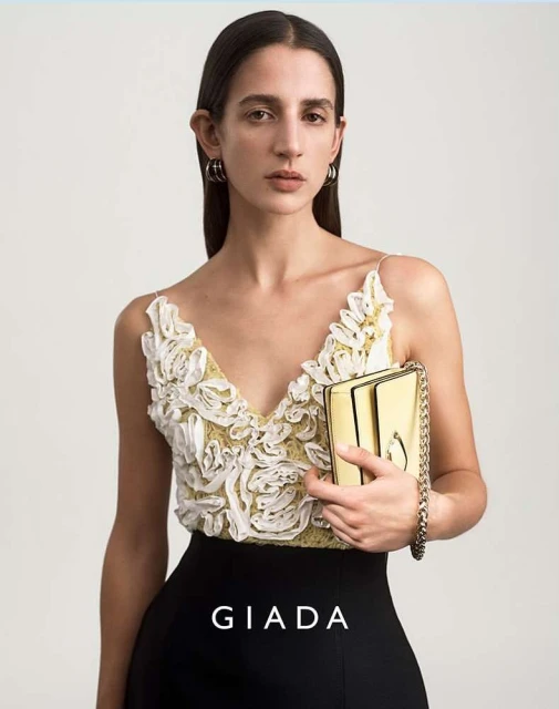 Fashion advertisement featuring a woman holding a gold clutch. The model wears a floral top and a black skirt in a neutral background.