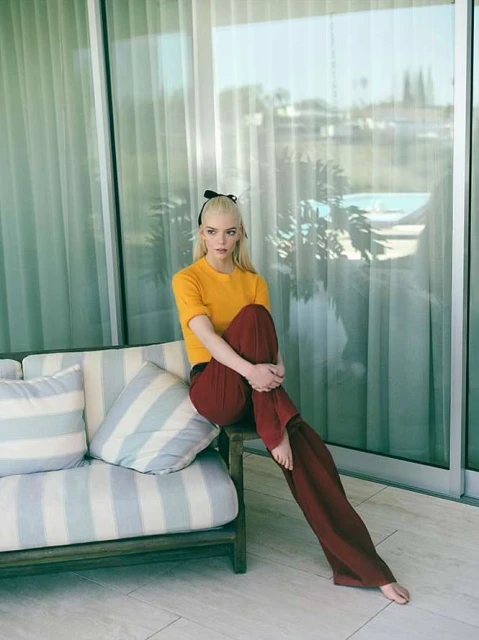 Woman with platinum blond hair sits on a striped couch wearing yellow top and wide burgundy pants. Balcony setting with pool view.