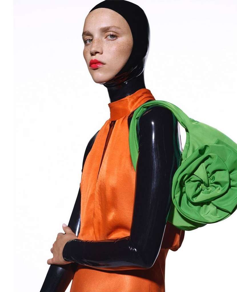 Fashionable woman poses in orange dress with green purse, black head covering, and arm coverings against a bright white backdrop.