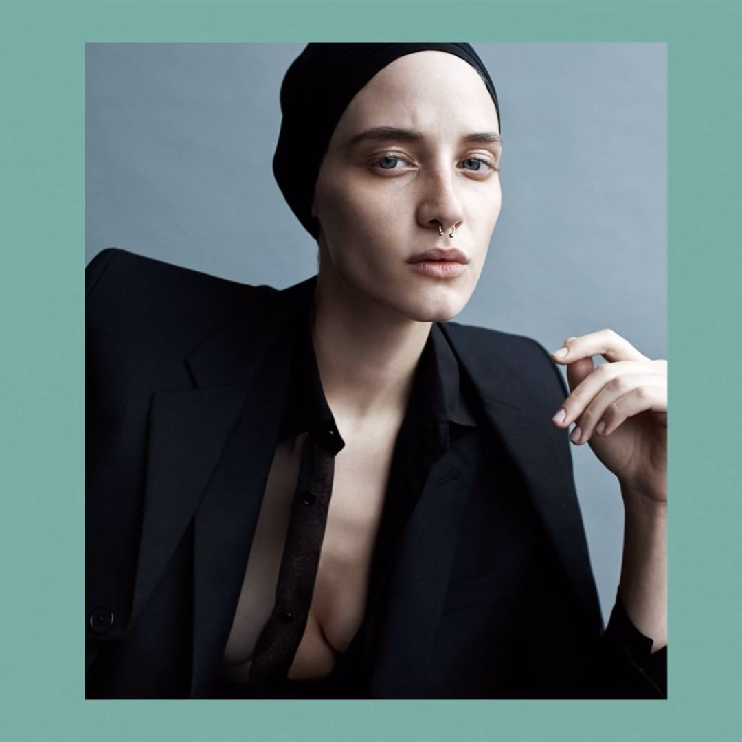 Close-up portrait of a young adult female with a nose ring, wearing a black head covering and jacket against a gray background.