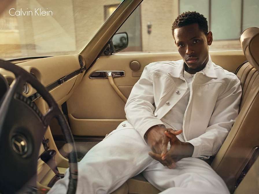 Black man wearing a white jacket and pants seated inside a vintage car with tan leather interior by photographer Josh Olins.