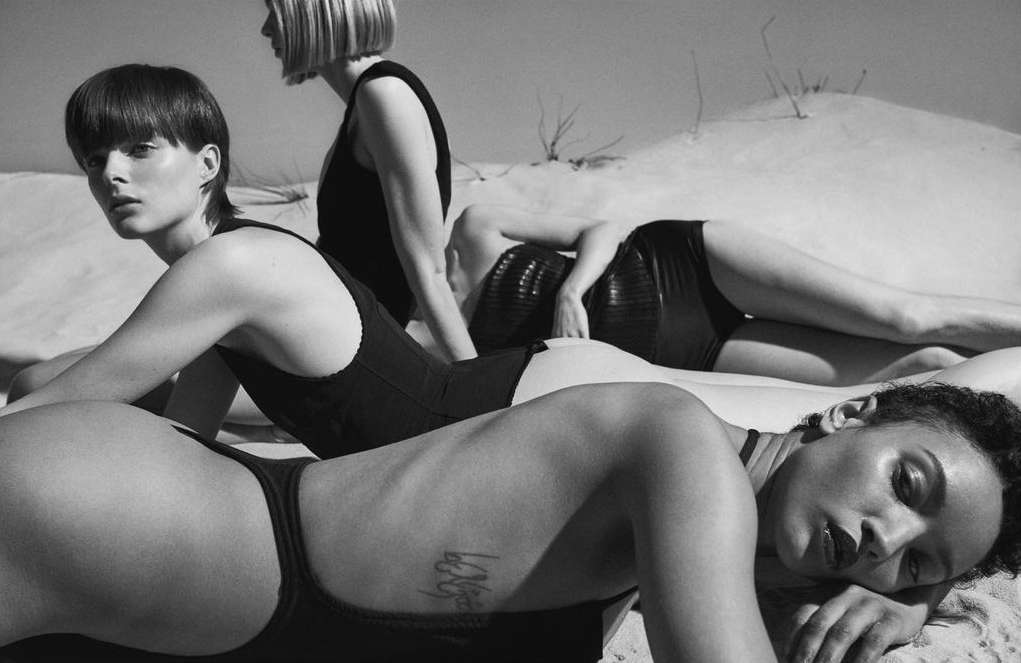 Three diverse women in black swimwear lying on sand, showcasing minimalist fashion and beauty against a natural, monochromatic backdrop.