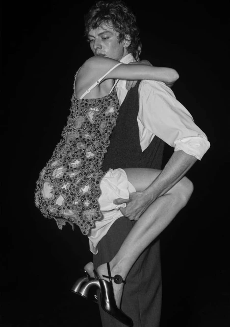 Monochrome portrait of a man holding a woman in his arms; she wears a patterned dress and heels, against a dark background.