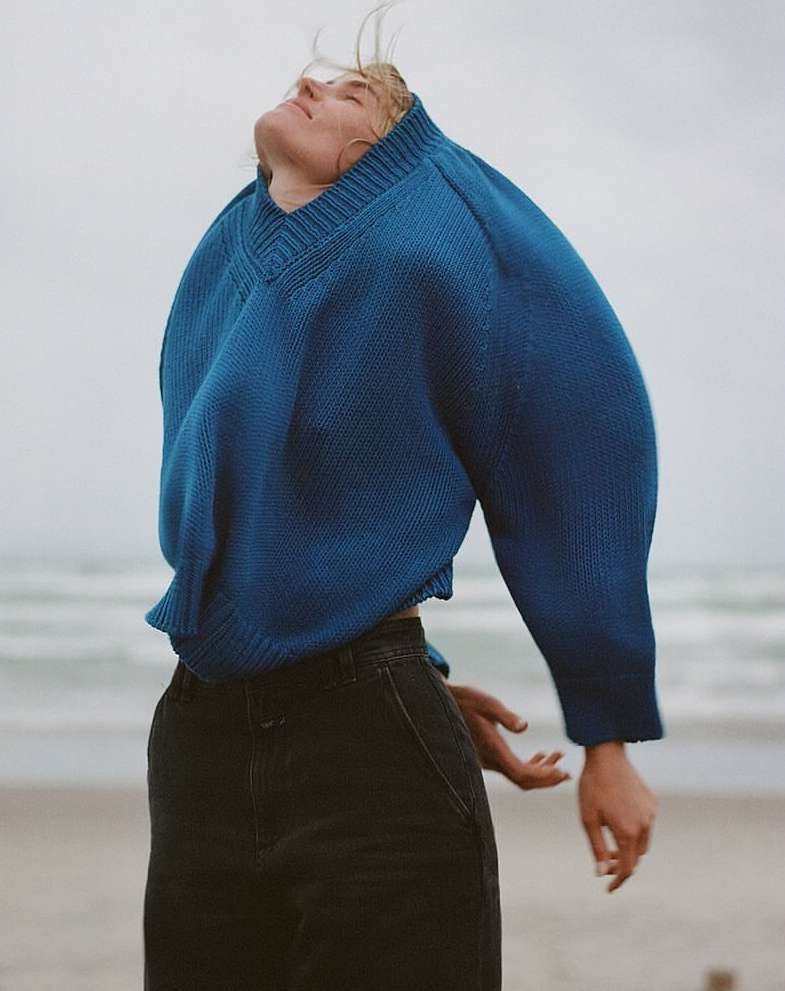 Woman in blue sweater and dark pants standing on a beach with her head tilted back toward the sky and soft light.