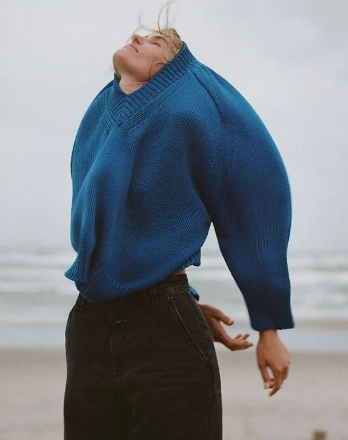 Woman in blue sweater and dark pants standing on a beach with her head tilted back toward the sky and soft light.