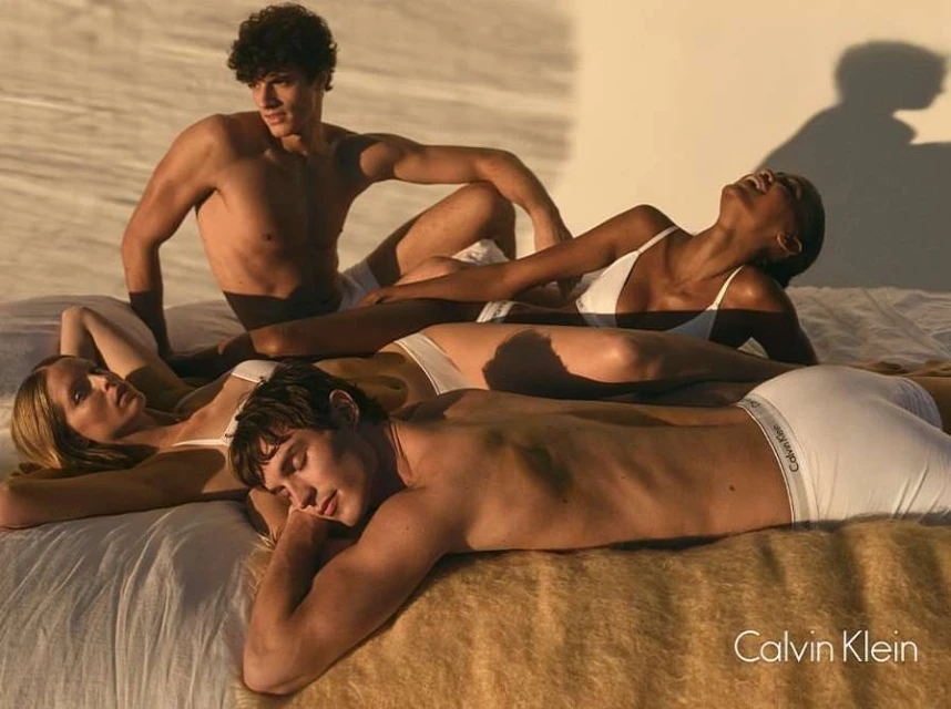 Four fashion models pose together on a bed in a sunlit photo studio, showcasing Calvin Klein underwear in a minimalist style.