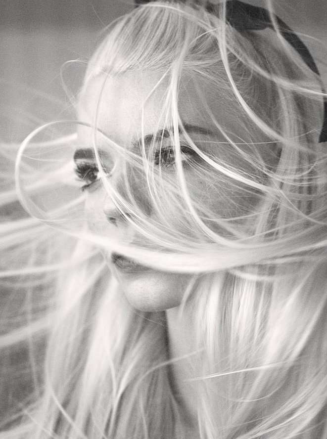 Monochrome portrait of a woman with long blonde hair blowing in the wind, ribbon in hair, freckles visible on face, close-up shot.