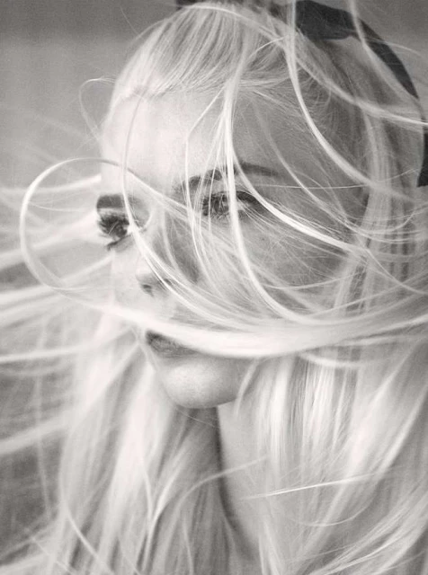 Monochrome portrait of a woman with long blonde hair blowing in the wind, ribbon in hair, freckles visible on face, close-up shot.