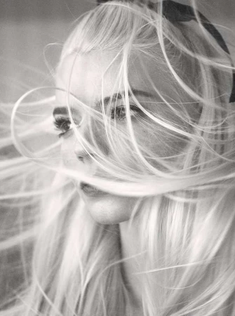 Monochrome portrait of a woman with long blonde hair blowing in the wind, ribbon in hair, freckles visible on face, close-up shot.