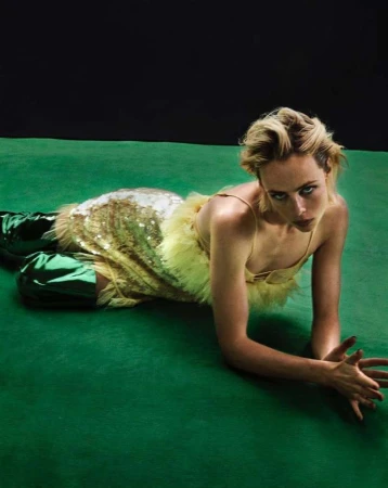 A woman with short blonde hair lies on a green surface, posing with her arms out in front of her. She wears a shiny gold dress.