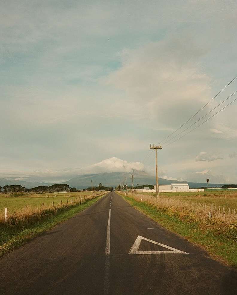 Scenic view of an empty asphalt road leading towards a distant mountain under a cloudy sky, framed by grassy fields.