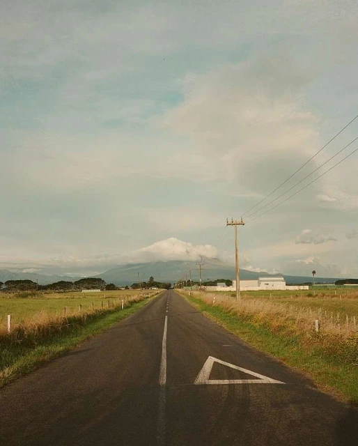 Scenic view of an empty asphalt road leading towards a distant mountain under a cloudy sky, framed by grassy fields.