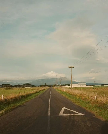 Scenic view of an empty asphalt road leading towards a distant mountain under a cloudy sky, framed by grassy fields.