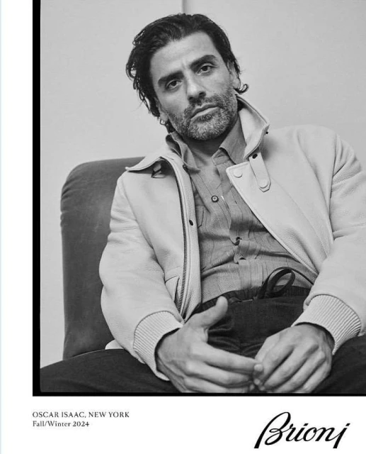 Black and white portrait of Oscar Isaac in an off-white jacket and button-down shirt, seated in a studio setting for Brioni.