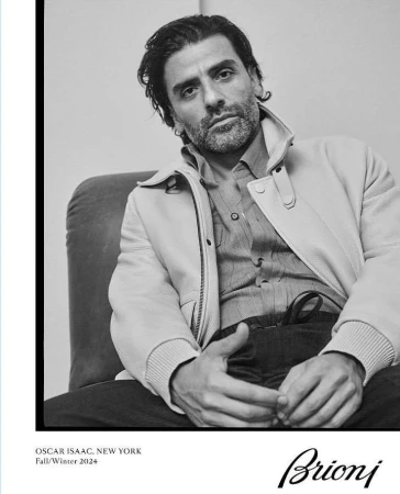 Black and white portrait of Oscar Isaac in an off-white jacket and button-down shirt, seated in a studio setting for Brioni.