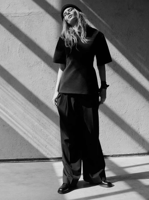 Black and white portrait of an adult woman posing in black clothing and a black beanie against a concrete wall with sunlight shadows.