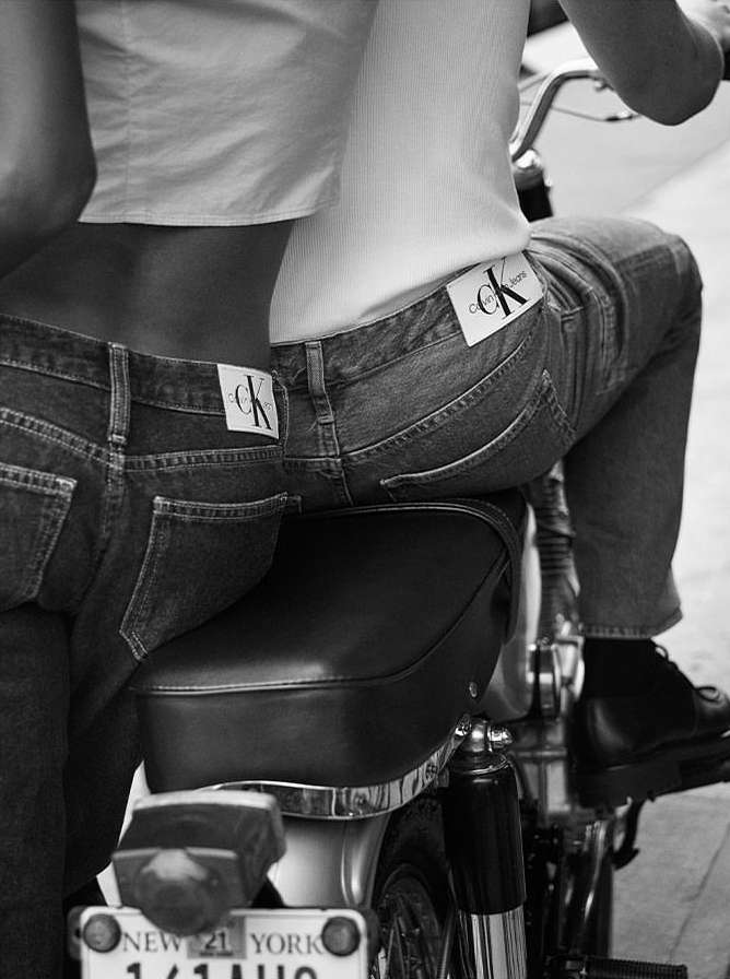 Close-up studio shot of two people on a motorcycle. Both wearing denim jeans with Calvin Klein logo. Black and white photography.