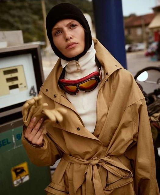 Stylish adult female model wearing ski goggles and a tan trench coat at a gas station in an outdoor daytime fashion shoot.
