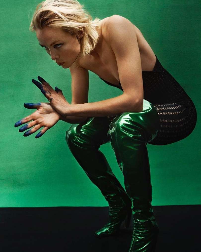Adult woman squats wearing a black top and shiny green thigh-high boots against a bright green backdrop, her hands painted blue.