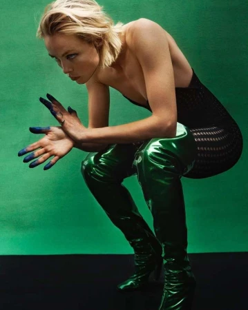 Adult woman squats wearing a black top and shiny green thigh-high boots against a bright green backdrop, her hands painted blue.