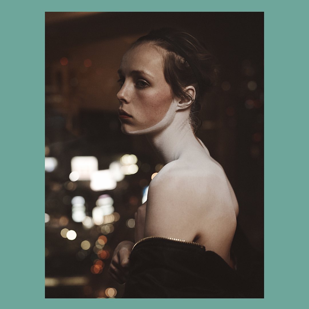 Close-up of a fair-skinned woman gazing left, partially undressed, with blurred city lights in the dark background. Low-key studio portrait.