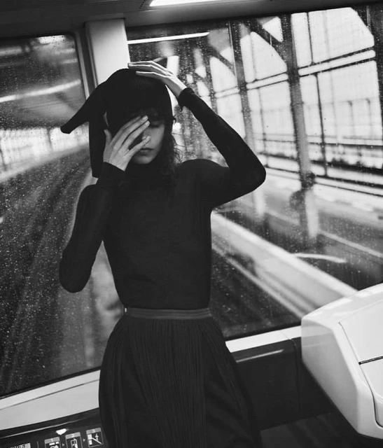 Black and white portrait of a woman in a train, wearing a turtleneck and a hat, with hands near her face, and a rainy window.