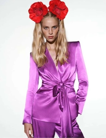 A fair-skinned woman poses in a purple satin suit with red flower headbands, against a bright white backdrop in a fashion shot.