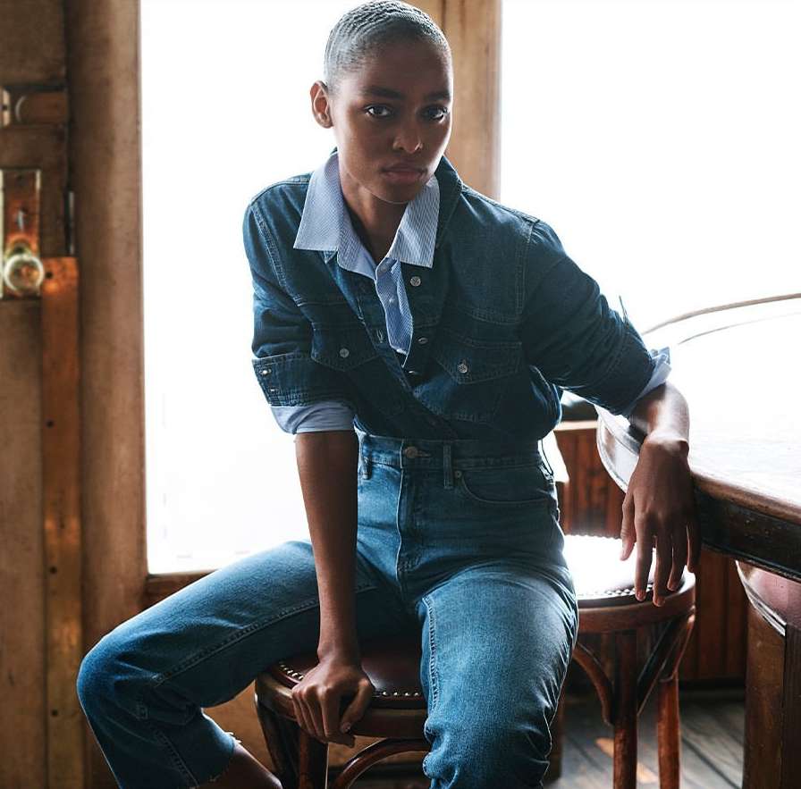 Model with short hair wearing a denim jacket and blue jeans seated on a wooden stool indoors