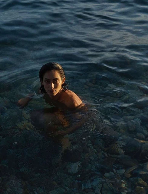 Woman emerges from the ocean with wet dark hair, looking at the camera with a serene expression, ocean water is a deep blue.