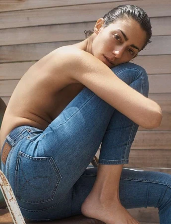 Woman wearing blue jeans posing seated outdoors against a wooden slat wall captured by Josh Olins