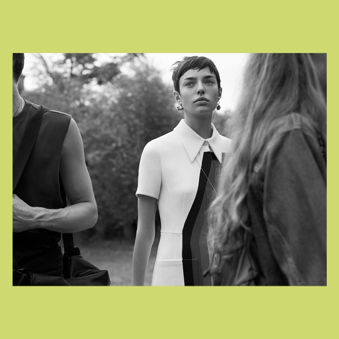 Black and white editorial photograph by Josh Olins of a short-haired woman in a white dress with a pointed collar, surrounded by figures outdoors.