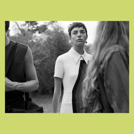 Black and white editorial photograph by Josh Olins of a short-haired woman in a white dress with a pointed collar, surrounded by figures outdoors.