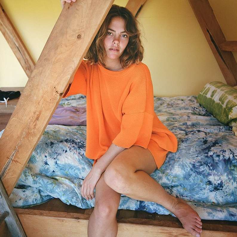 A woman with shoulder-length hair wears an orange sweater and sits on a patterned bed in a room with wooden beams, posing for a portrait.