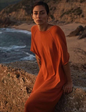 Adult woman in an orange dress poses outdoors on a stone wall with the ocean and rocky hills in the background during daytime.