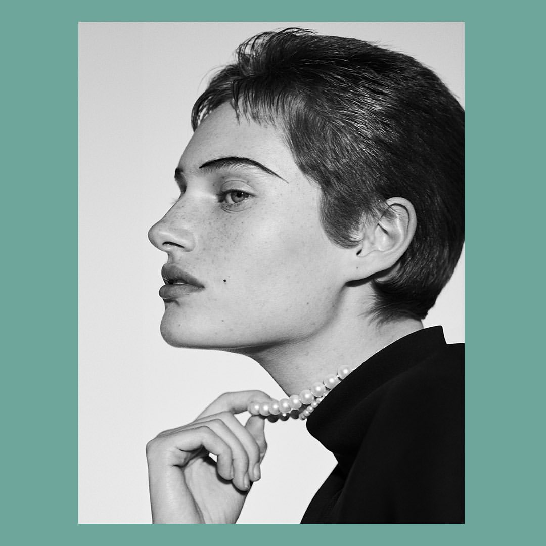 Black and white profile portrait of a woman with short hair wearing a pearl necklace and dark top by Josh Olins
