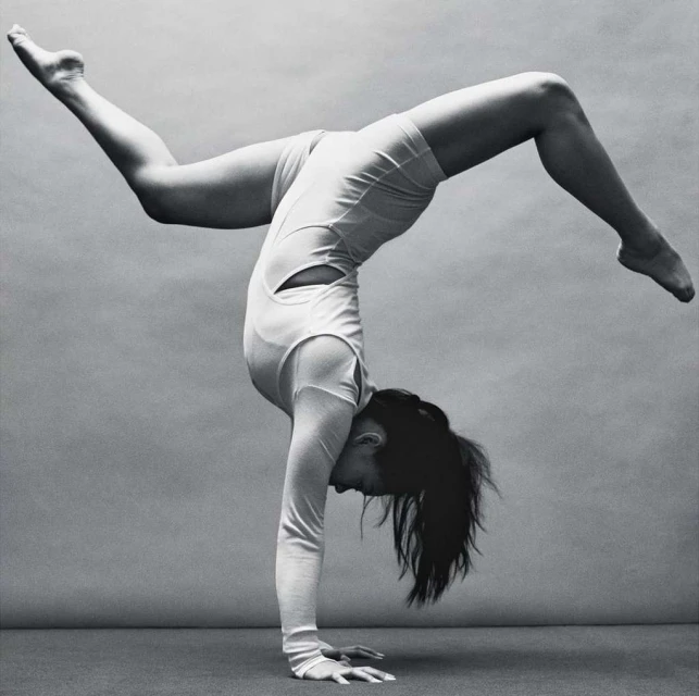 Athletic woman in a white bodysuit performing a handstand against a grey backdrop in a fitness studio. Demonstrating balance and flexibility.