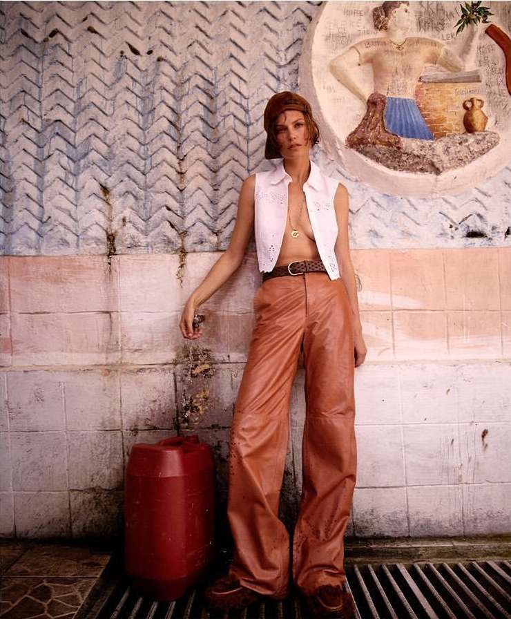 An adult female model leans against a wall in an outdoor setting, wearing a white top and brown leather pants, next to a red container.