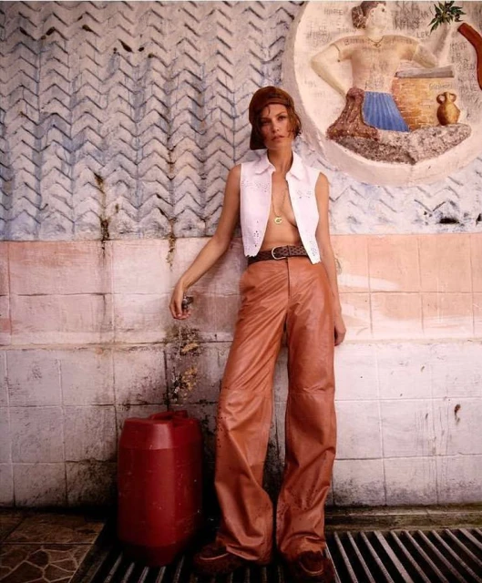 An adult female model leans against a wall in an outdoor setting, wearing a white top and brown leather pants, next to a red container.